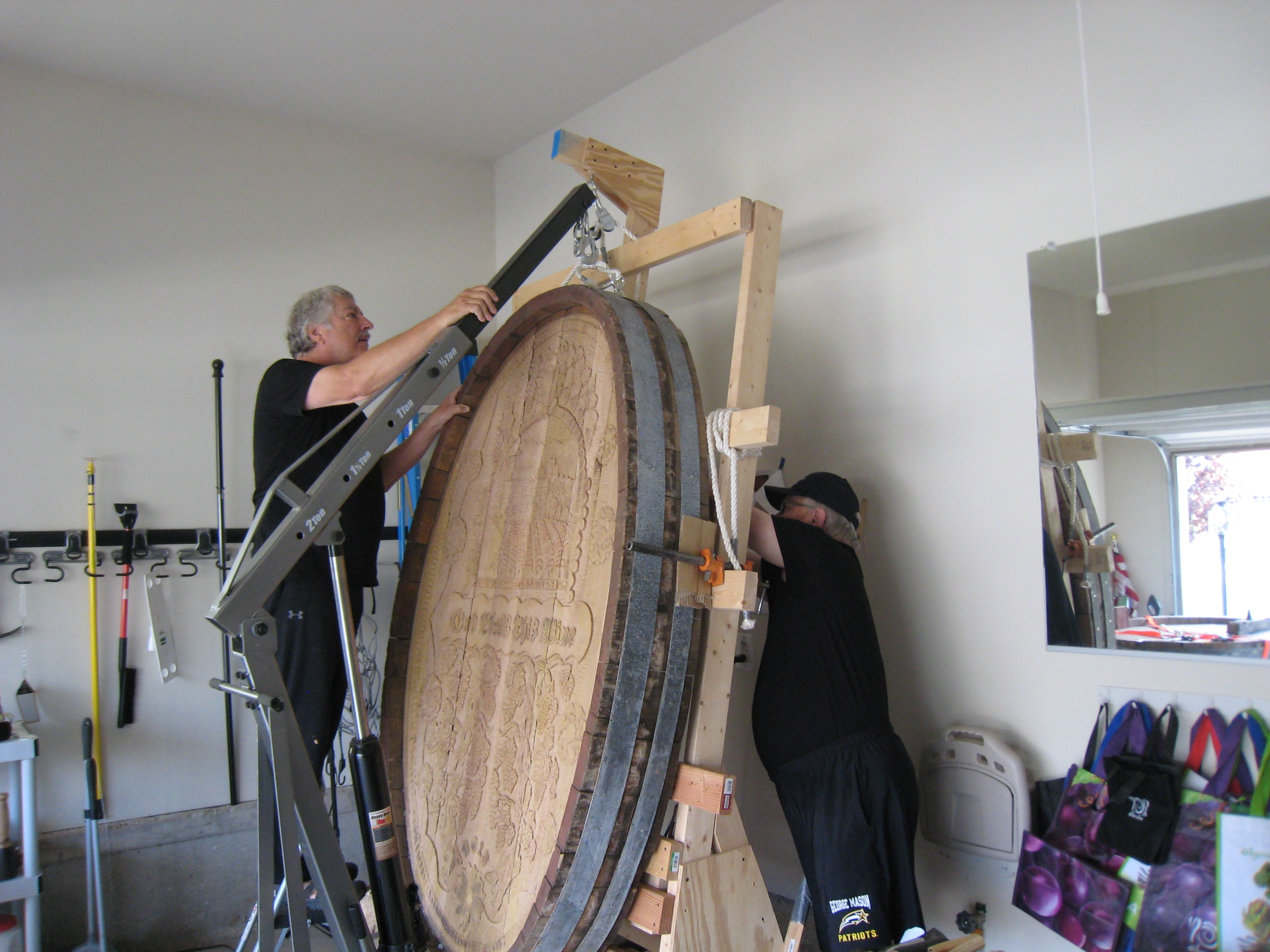 Two people using the garage crane and rigging to reposition the massive carved cask end on the carving rack